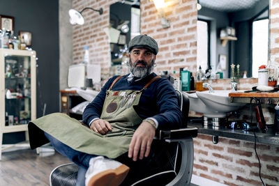 Middle aged male barber in cap and apron sitting on chair in grooming salon and looking at camera