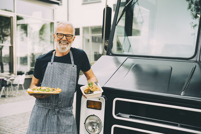 Smiling senior owner with food plate standing against commercial land vehicle