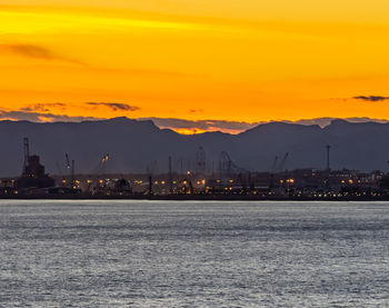 Illuminated city by sea against orange sky