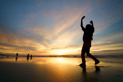 Low angle view of silhouette man with arms raised walking at beach during sunset