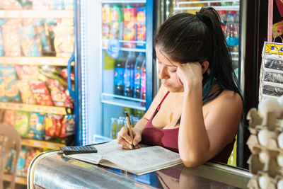 Latina woman very thoughtful while writing down in a notebook the accounts of her grocery store.