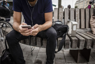 Midsection of man using mobile phone while sitting on wooden bench in city