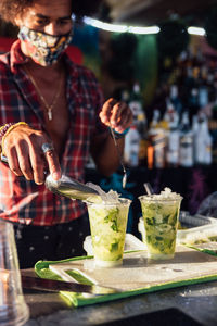 Black male bartender in protective mask adding ice into glass with lime while preparing tropical cocktail at counter of beach bar