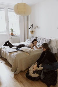 Smiling woman unpacking luggage near female friend lying down on bed in apartment