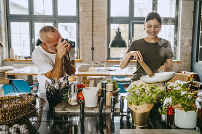 Happy female chef cooking food with male colleague photographing her in studio