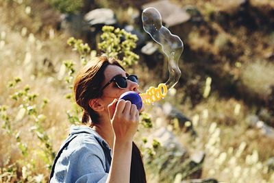 Side view of young woman blowing bubble while standing against plants