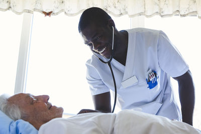 Happy male nurse examining senior man with stethoscope in hospital ward