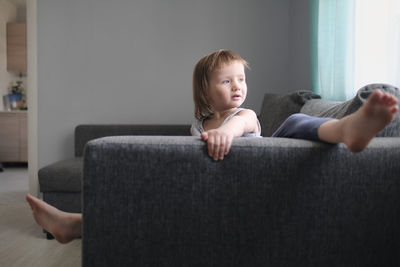 European toddler child sits and gets bored on gray sofa in bright living room, minimalist interior