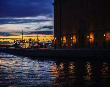 Illuminated buildings by river against sky at sunset