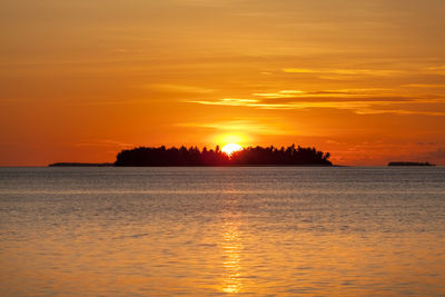 View of tiny tropical island and sun going down behind it, colouring orange all around maldives