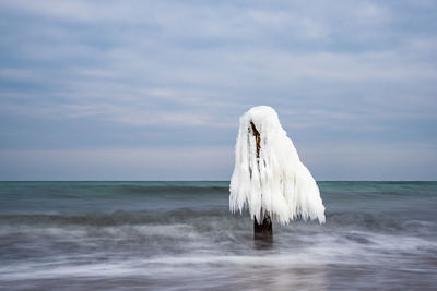 Frozen wooden post in sea against cloudy sky