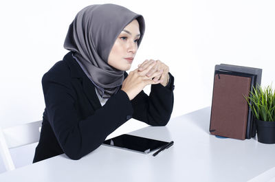 Young woman looking away while sitting on table against white background