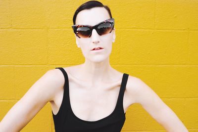 Portrait of young woman wearing sunglasses standing against yellow wall