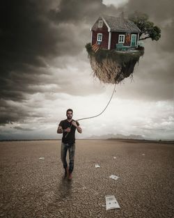 Digital composite image of young man holding house with rope on field against cloudy sky