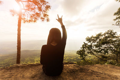 Rear view of woman making horn gesture while sitting on cliff against sky