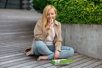 Young beautiful blond woman eating sushi outdoors, on the wooden terrace, by modern building