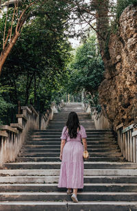 Rear view of young woman wearing pink dress, walking up long stairs in park in city