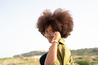 Side view of smiling young african american female with curly hair touching glasses and looking at camera while enjoying summer holidays in countryside