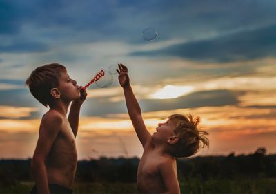 Shirtless friends playing with bubbles against sky during sunset
