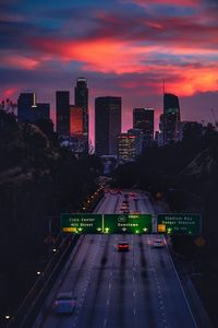 Illuminated road amidst buildings in city against sky during sunset