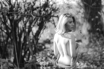 Rear view of topless young woman looking away while standing against trees