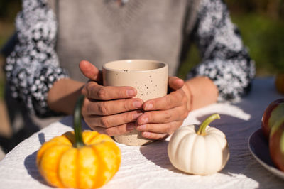 The working hands of the girl hold a ceramic cup standing on the table near small pumpkins