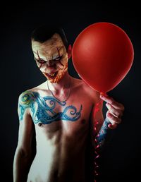 Scary shirtless man with face paint holding red balloon against black background