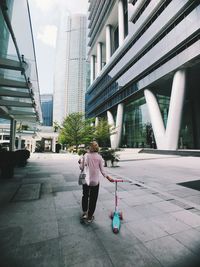 Rear view of woman with push scooter walking on footpath against buildings