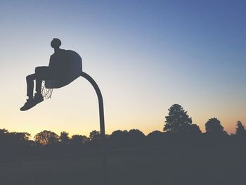 Low angle view of silhouette man sitting on basketball hoop against clear sky