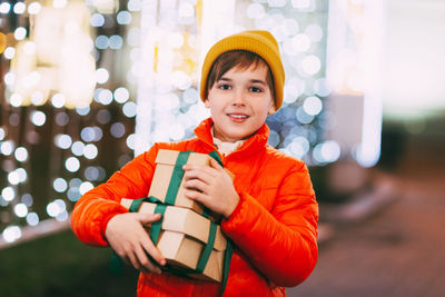 A happy boy in an orange jacket with a gift box in his hands at a christmas market on a winter