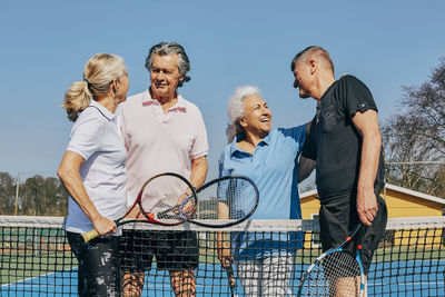 Happy male and female seniors greeting while standing at tennis court against clear sky