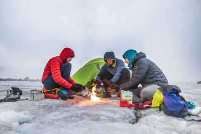 Three climbers warm their hands over a fire while camping on a glacier