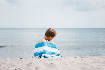 Young boy sat wrapped in a striped towel alone on the beach