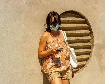 Woman using phone on a bus stop wearing surgical protective mask due to the coronavirus normative
