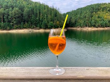 Glass of aperol spritz cocktail with lake and green forest in the background