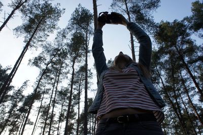 Low angle view of woman photographing through camera while standing against trees at forest