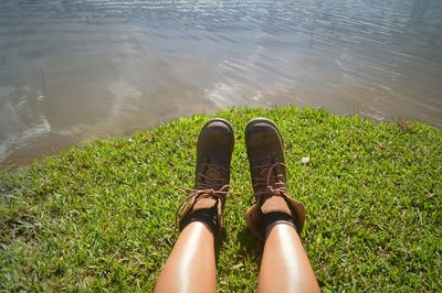 Low section of woman wearing brown shoes sitting on grassy field by lake during sunny day