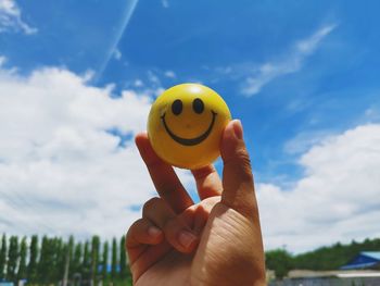Cropped hand holding ball with anthropomorphic smiley face against sky