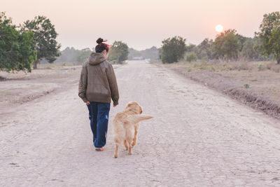 Rear view of woman walking by dog on road during sunrise