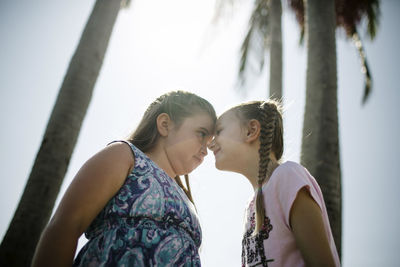 Low angle view of sisters touching foreheads while standing by coconut palm trees at beach against clear sky during sunny day