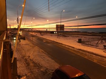 Road by illuminated street against sky during sunset