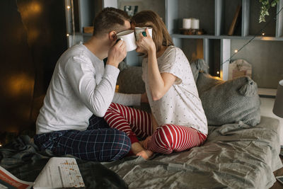 Side view of couple holding mugs while sitting on bed at home