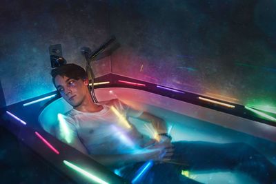 Digital composite image young man looking away while lying in illuminated multi colored bathtub