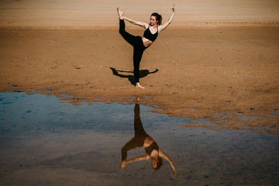 Side view of flexible female standing on sandy shore of river in natarajasana and practicing yoga while balancing on leg with eyes closed