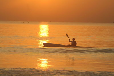 Silhouette man canoeing in sea against orange sky during sunrise