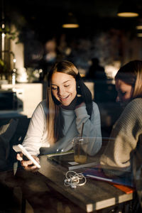 Smiling teenage girl showing smart phone to friend while sitting in cafe