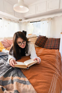 Woman with eyeglasses reading book while lying on bed at home