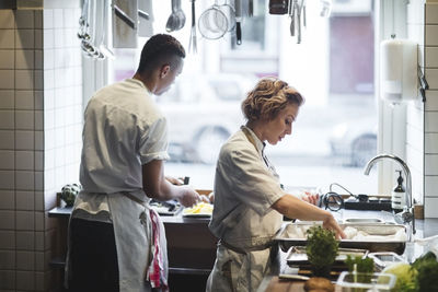 Side view of female chef preparing food on counter in kitchen with male colleague working at restaurant