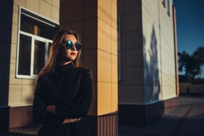 Young woman wearing sunglasses looking away while standing against built structure