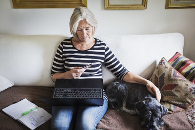 Senior woman using credit card and laptop while stroking dog on sofa